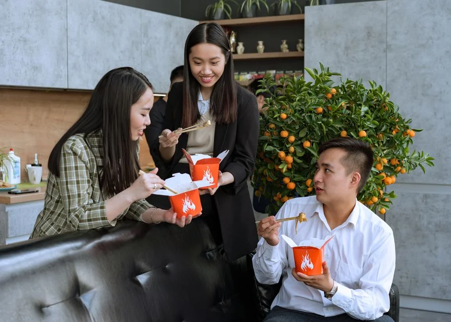 Group of colleagues enjoying a break eating takeout food with chopsticks indoors.